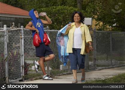 Mature woman walking with a teenage boy leaning on a chain-link fence and drinking water beside her