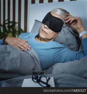 Mature woman wearing black sleeping mask, lying in bed in bedroom. Mature Woman Wearing Black Sleep Mask, Lying in Bed