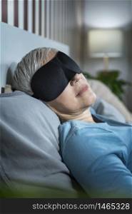 Mature woman wearing black sleeping mask, lying in bed in bedroom. Mature Woman Wearing Black Sleep Mask, Lying in Bed