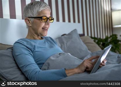 Mature woman wearing blue light blocking glasses with amber lenses, lying in bed before sleep, looking at tablet screen . Mature Woman Wearing Blue Light Blocking Eye Glasses, Using Tablet