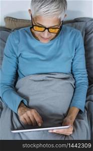 Mature woman wearing blue light blocking glasses with amber lenses, lying in bed before sleep, looking at tablet screen . Mature Woman Wearing Blue Light Blocking Eye Glasses, Using Tablet