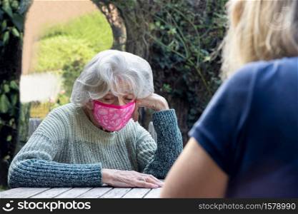 Mature Woman Wearing Mask Visiting Lonely Depressed Senior Mother In Garden During Lockdown
