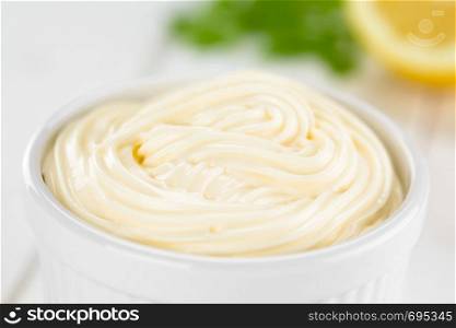 Mayonnaise sauce in white bowl with lemon and parsley in the back (Selective Focus, Focus in the middle of the image). Mayonnaise