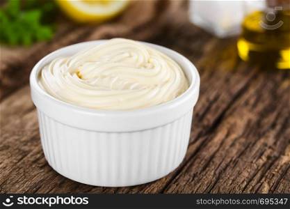 Mayonnaise sauce in white bowl with parsley, lemon, salt and oil in the back (Selective Focus, Focus in the middle of the mayonnaise). Mayonnaise