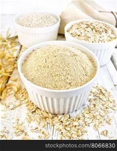 Meal oat, bran and oat flakes in three white bowls, oat stalks, towel on a background of wooden boards