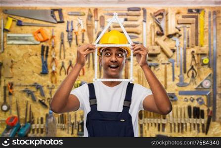 measurement, construction and building - happy smiling indian worker or builder in helmet with folding ruler in shape of home over working tools on wall at workshop background. happy indian builder with ruler in shape of home