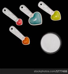 Measuring spoons with heart shaped bowls pointing to a round bowl of sugar on a black background.