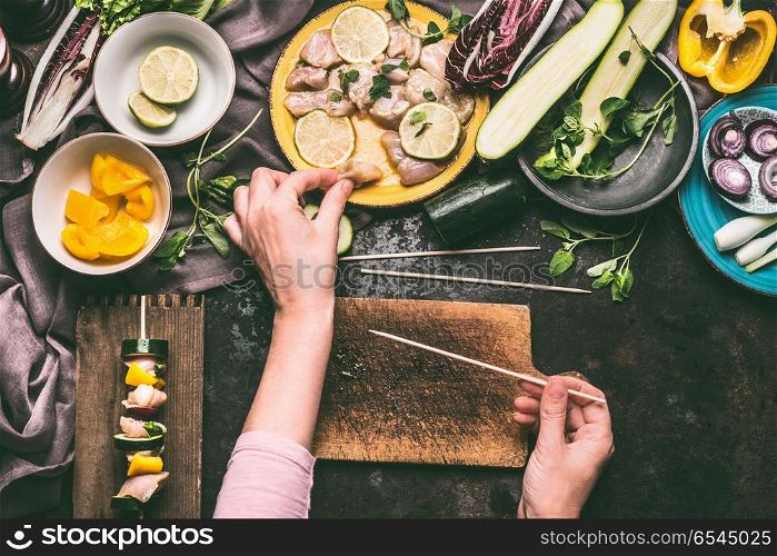 Meat and vegetables skewers preparation. Female hand takes a piece of chicken on dark kitchen table background with bowls with ingredients for grill, top view