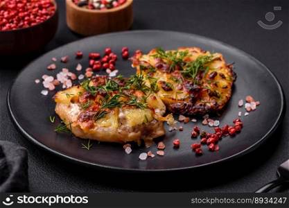 Meat baked in the oven with pineapple, tomatoes, cheese, spices and herbs on a black ceramic plate on a dark concrete background