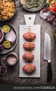 Meat balls cooking. Raw meat balls on white cutting board with knife on kitchen table background with rice and salad dishes, top view
