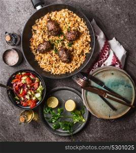Meat balls with vegetables rice in pan and salad, served on gray stone kitchen table background with plates and cutlery, top view