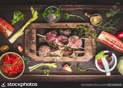 Meat skewers in wooden plate with grill and cooking ingredients on dark rustic background, top view