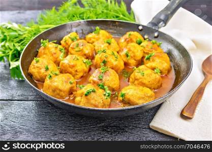 Meatballs with tomato sauce in a frying pan, parsley, dill, napkin and spoon on a black wooden board background