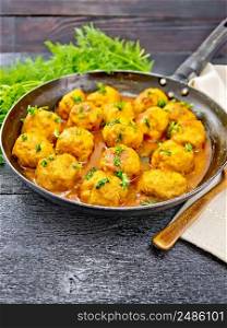 Meatballs with tomato sauce in a frying pan with parsley greens, dill, towel and spoon on a black wooden board background