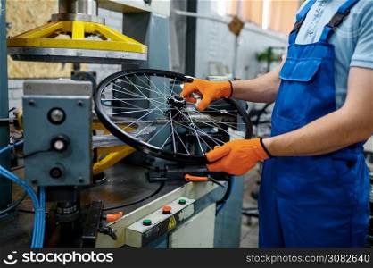 Mechanic in uniform holds bicycle wheel near machine tool on factory. Bike rims and spokes assembly in workshop, cycle parts installation, modern technology. Mechanic holds bicycle wheel near machine tool