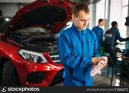 Mechanic wipes his hands after repairing the car, motor diagnostic. Vehicle with opened hood on background, auto-service. Mechanic wipes his hands after repairing the car