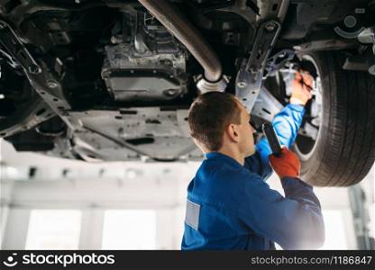 Mechanic with lamp checks car brake hoses, repair station. Tire service, vehicle maintenance