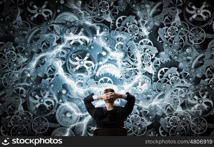 Mechanism of teamwork and collaboration. Young relaxed businessman sitting in chair and looking at gears mechanism