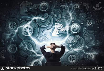 Mechanism of teamwork and collaboration. Young relaxed businessman sitting in chair and looking at gears mechanism