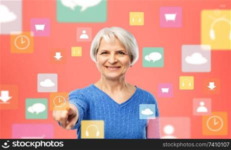 media, technology and old people concept - portrait of smiling senior woman in blue sweater pointing finger to mobile app clock icon over living coral background. senior woman pointing to mobile app clock icon