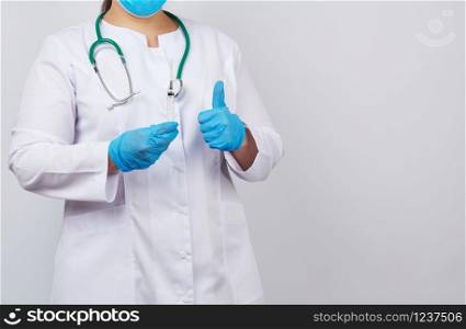 medic woman in white coat and blue latex gloves holds a syringe and shows like gesture with hand, white studio background, concept of timely vaccination against viruses, copy space