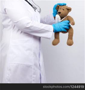 medic woman in white coat with buttons holding a brown teddy bear with a white gauze bandage tied with her paw, concept of pediatrics and animal treatment