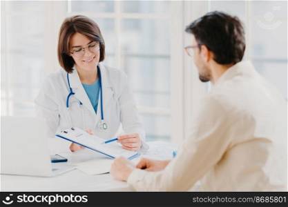 Medical appointment. Female doctor gives professional medical help to male patient, explains written information on paper in clipboard, gives support and good service, pose at hospital near desktop.