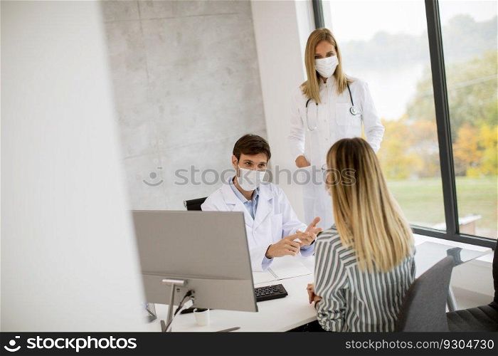 Medical couple doctors with protective medical facial masks talking with young female patient and using computer in the offic