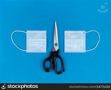 Medical disposable face mask cut in half and scissors on blue background.. Medical disposable face mask cut in half and scissors on a blue background.