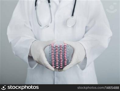 Medical doctor wearing clear latex gloves holding stack of different pills, antibiotics and virus treatment tablets on grey hospital wall.