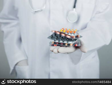 Medical doctor wearing clear latex gloves holding stack of different pills on grey hospital wall. Antibiotics and virus treatment tablets.