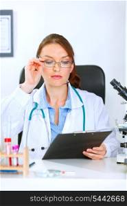 Medical doctor woman sitting at office table and looking in clipboard&#xA;