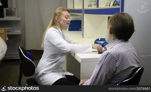 Medical exam. Confident smiling female doctor in lab coat checking senior man&acute;s temperature with electronic thermometer in doctor&acute;s office during checkup. Sick old man measuring body temperature during visit a hospital. Slow motion.