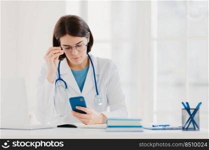 Medical technology concept. Concentrated woman doctor looks at smartphone device, checks necessary information in internet, wears stethoscope around neck, big round glasses, poses in clinic.