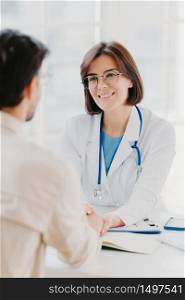 Medical trust concept. Vertical image of female doctor reassures her male patient, hold hand and talk calmly about illness cure, gives professional consultation and support, pose at doctors office