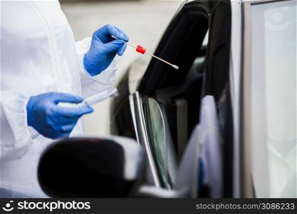 Medical worker in N95 PPE performing nasal & throat swab on person in vehicle through car window,COVID-19 UK NHS mobile testing centre drive thru facility,hands closeup in blue gloves holding test kit
