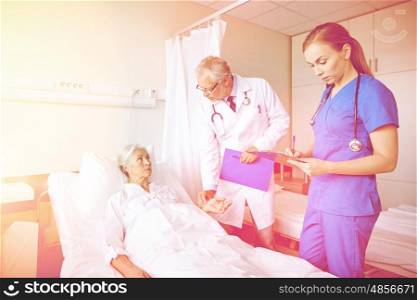 medicine, age, health care and people concept - doctor and nurse with clipboards visiting senior patient woman and checking her pulse at hospital ward