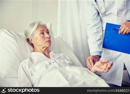 medicine, age, health care and people concept - doctor with clipboard visiting senior patient woman and checking her pulse at hospital ward