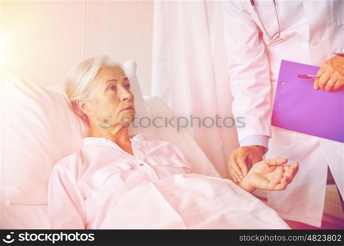 medicine, age, health care and people concept - doctor with clipboard visiting senior patient woman and checking her pulse at hospital ward