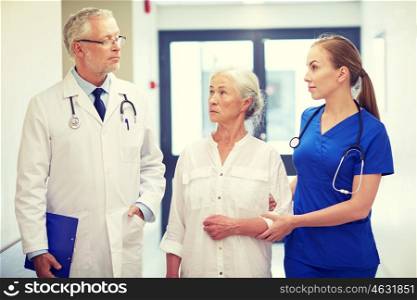 medicine, age, health care and people concept - male doctor with clipboard, young nurse and senior woman patient talking at hospital corridor