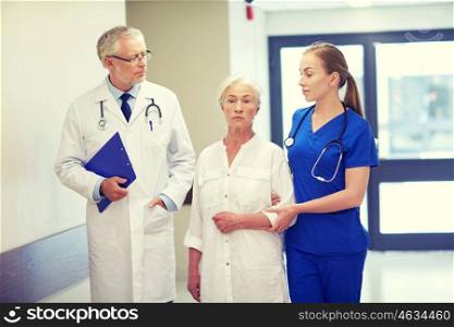 medicine, age, health care and people concept - male doctor with clipboard, young nurse and senior woman patient going down corridor at hospital