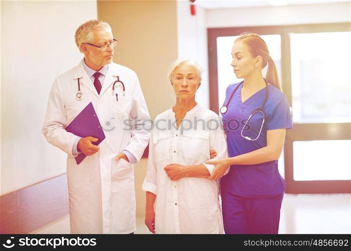 medicine, age, health care and people concept - male doctor with clipboard, young nurse and senior woman patient going down corridor at hospital