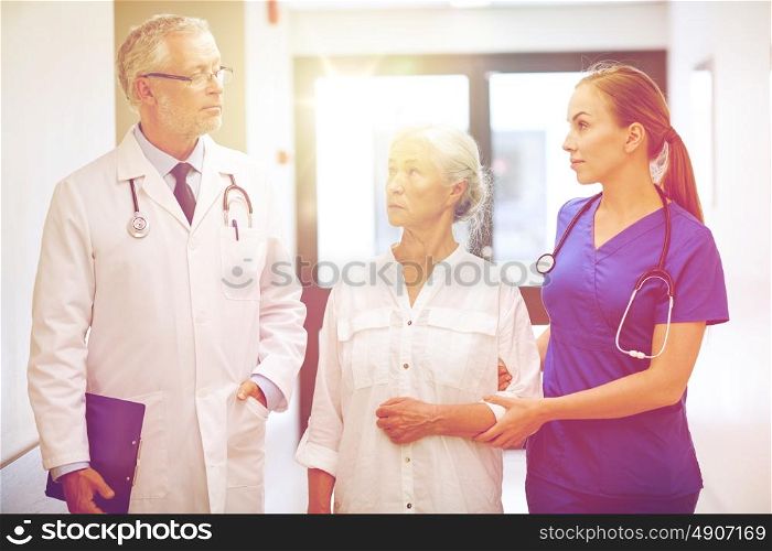 medicine, age, health care and people concept - male doctor with clipboard, young nurse and senior woman patient talking at hospital corridor. medics and senior patient woman at hospital