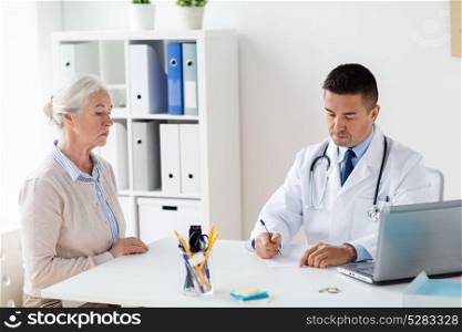 medicine, age, healthcare and people concept - senior woman and doctor with laptop computer writing prescription at hospital. woman and doctor with prescription at clinic