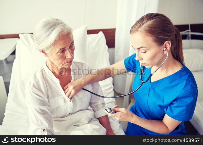 medicine, age, support, health care and people concept - doctor or nurse with stethoscope visiting senior woman and checking her heartbeat at hospital ward