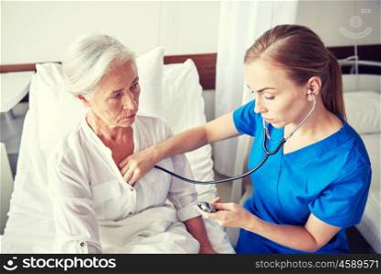 medicine, age, support, health care and people concept - doctor or nurse with stethoscope visiting senior woman and checking her heartbeat at hospital ward