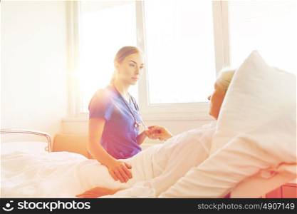 medicine, age, support, health care and people concept - doctor or nurse visiting and cheering senior woman lying in bed at hospital ward. doctor or nurse visiting senior woman at hospital