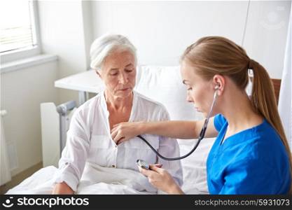 medicine, age, support, health care and people concept - doctor or nurse with stethoscope visiting senior woman and checking her breath or heartbeat at hospital ward