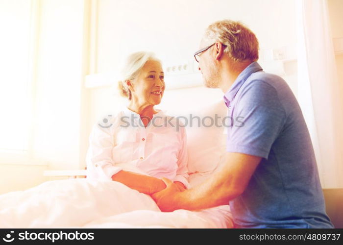 medicine, age, support, health care and people concept - happy senior man visiting and cheering his woman lying in bed at hospital ward