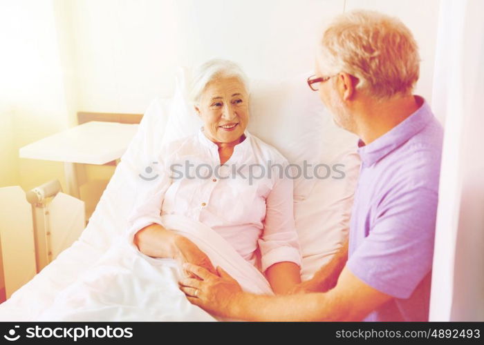 medicine, age, support, health care and people concept - happy senior man visiting and cheering his woman lying in bed at hospital ward